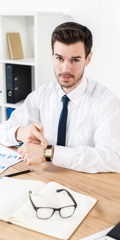 young-businessman-looking-camera-pointing-his-finger-toward-watch-workplace